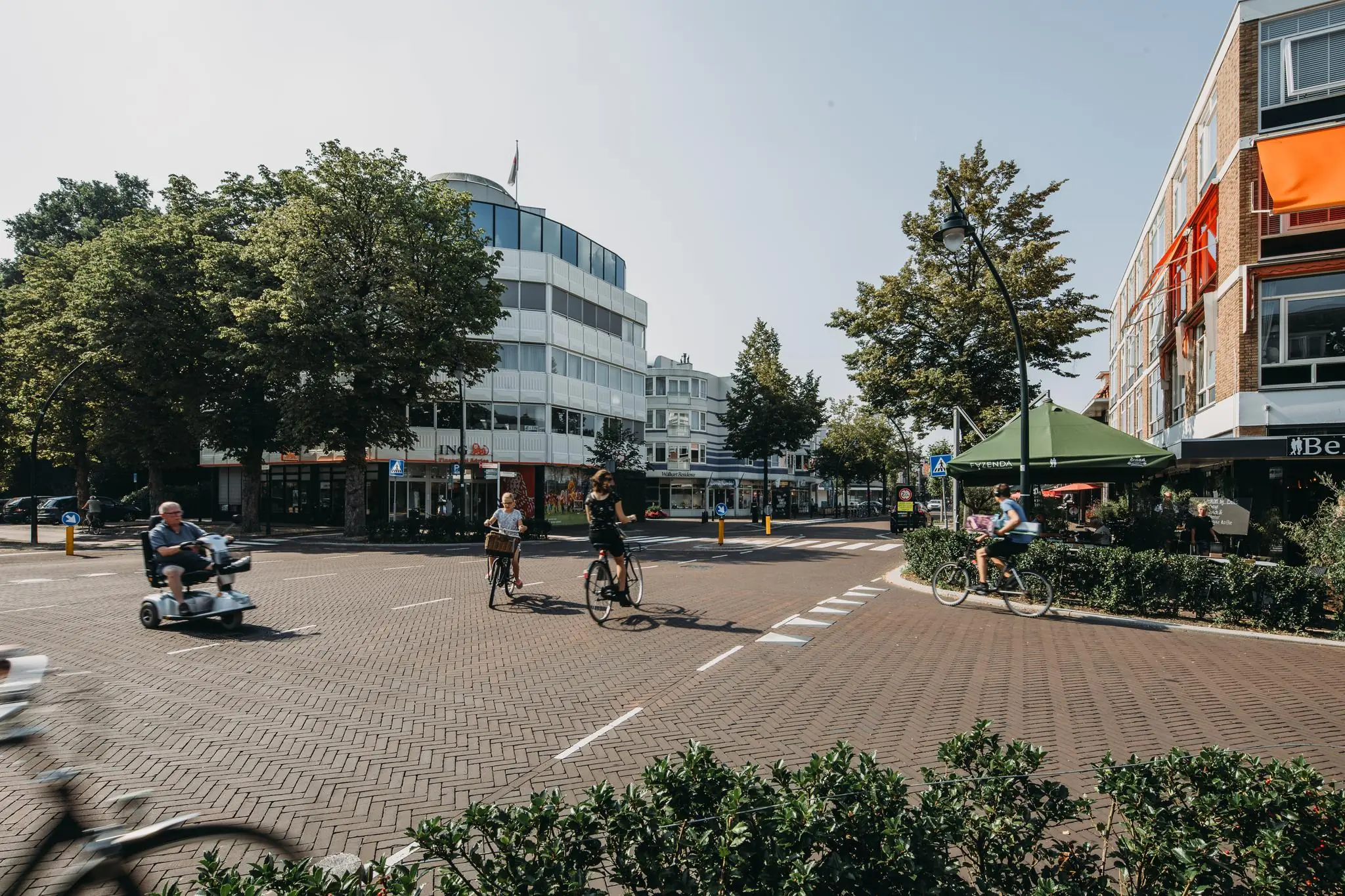 Verschillende mensen op fietsen en een scootmobiel rijden over een kruispunt op de 1e Hogeweg in een zonnige, stedelijke omgeving.